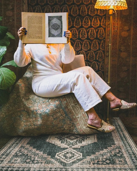 Person sitting on a patterned rug holding a book in front of their face in a cozy room.